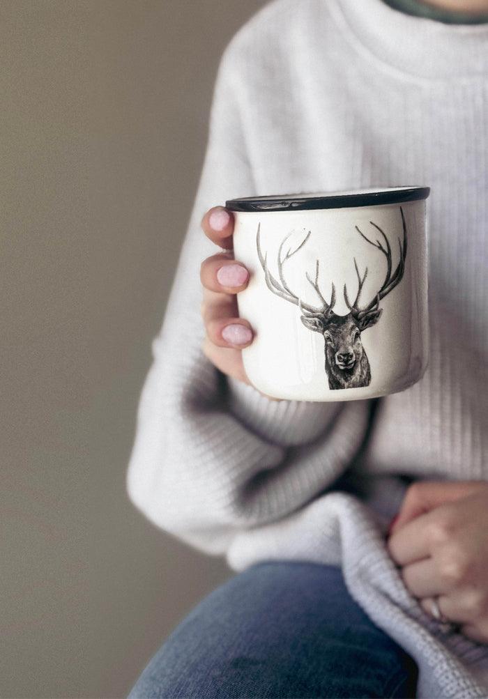 A person in a light gray sweater and jeans holds an Artsy Ems Designs enamel mug featuring a detailed deer with antlers, black rim, and light pink nails—perfect for nature lovers.