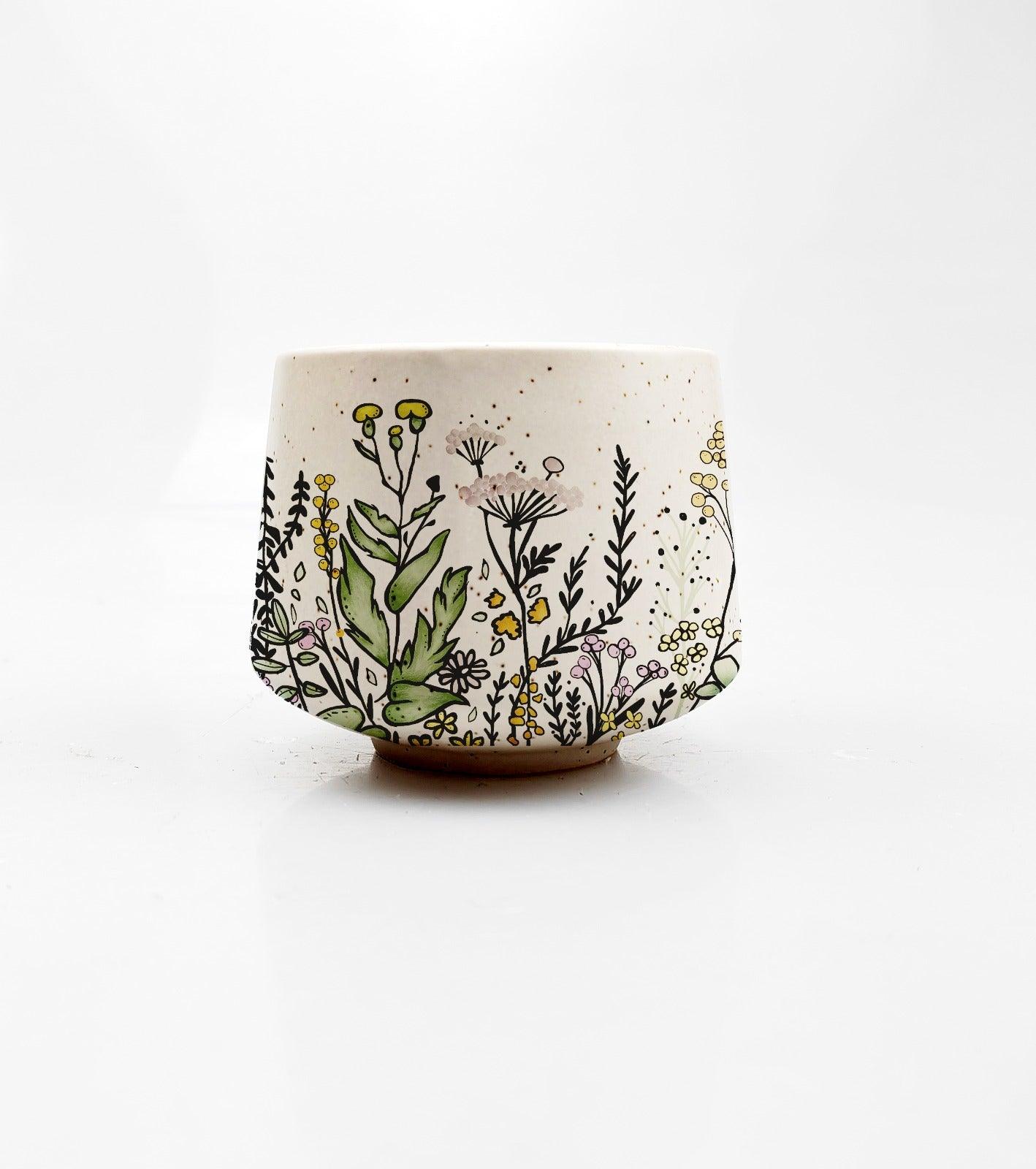 The Floral Bloom Mini Cup by Artsy Ems Designs is a white ceramic cup adorned with hand-painted wildflowers in yellow, purple, and white, plus green leaves, all showcasing exquisite craftsmanship against a plain white background.