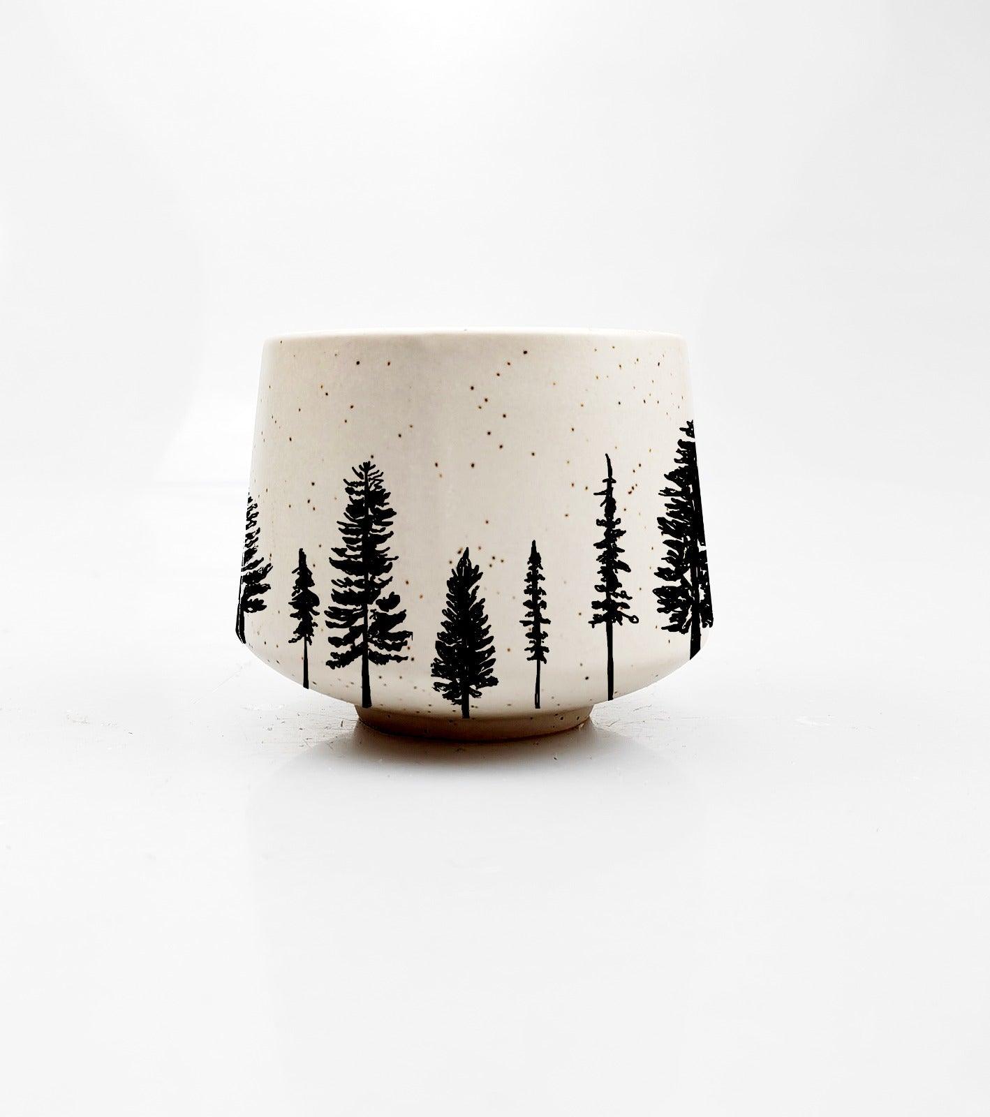 The Evergreen Elegance Mini Cup by Artsy Ems Designs is a small cup with black pine tree silhouettes around the base, brown speckles on a plain white background—a unique piece of drinkware.
