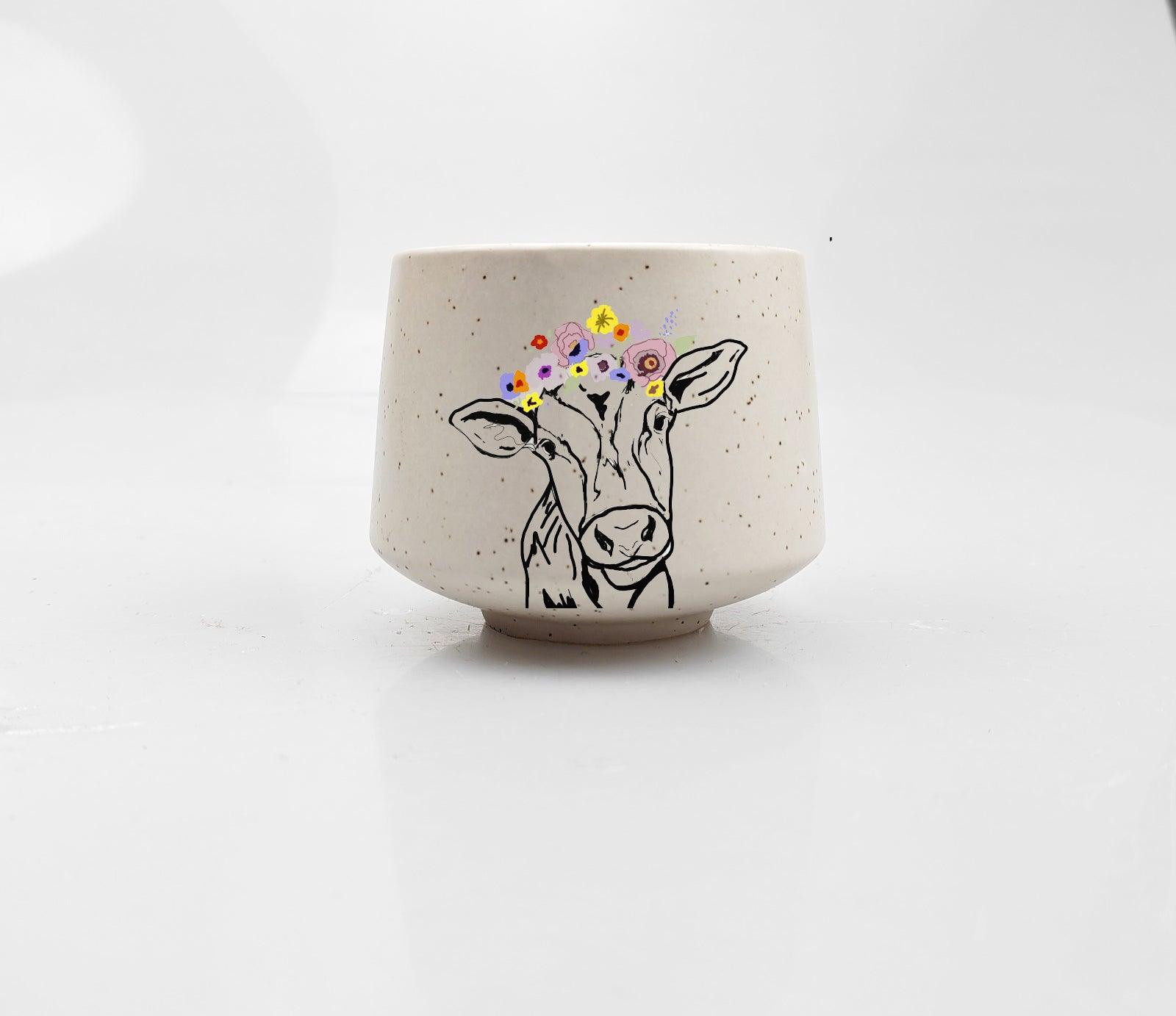 Artsy Ems Designs white ceramic Artisan Cow Cup features a handcrafted look with a black line cow wearing a colorful flower crown—perfect for whimsical home décor and displayed on a plain white surface.