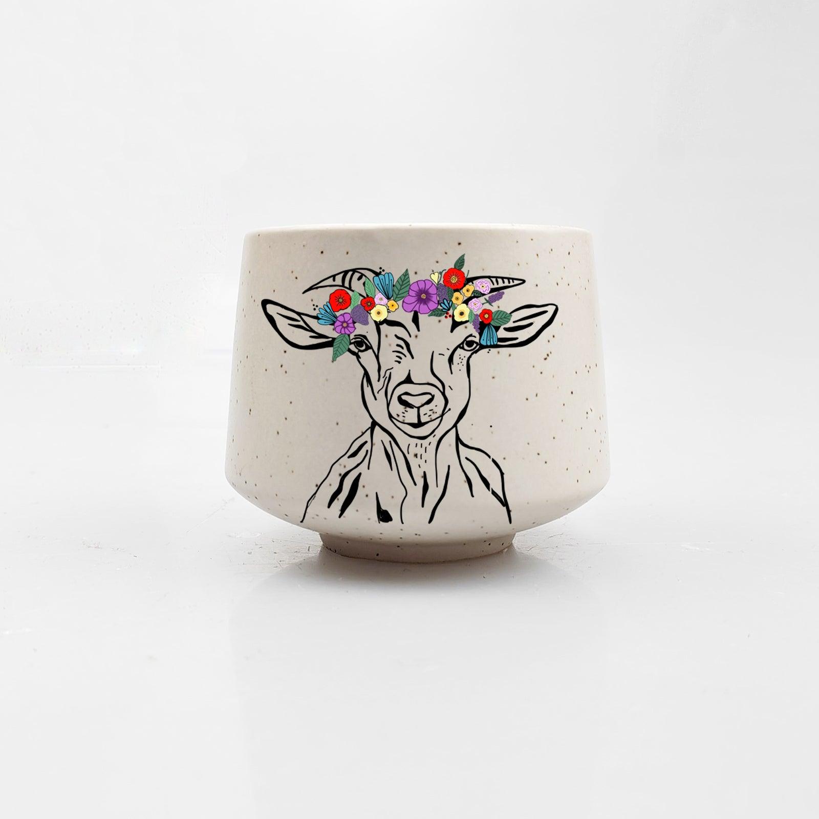 This white ceramic cup from Artsy Ems Designs showcases an artisan-drawn goat wearing a vibrant flower crown, set on a plain white background.